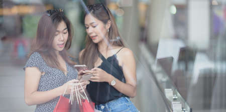 Two beautiful asian women chatting together and holding smartphone while shopping at the mallの写真素材