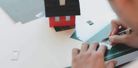 Close-up view of young male architect making small house model while cutting the cardboard with blade cutterの素材 [FY310130788153]