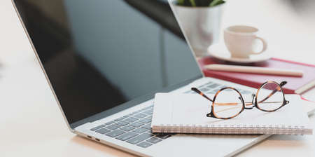 Cropped shot of cozy workplace with open laptop computer, glasses and office supplies on white wooden tableの写真素材