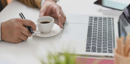 Close-up view of businessman holding a coffee cup while working on his project with laptop computerの写真素材