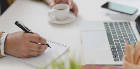 Close-up view of young businessman working on his project while writing the idea on notebook and drinking a cup of coffeeの写真素材