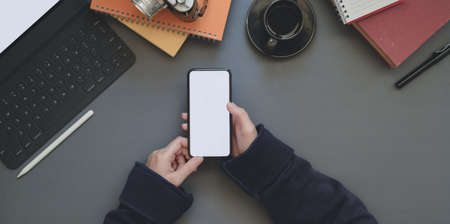 Overhead shot of young female holding blank screen smartphone in dark trendy workplace with office suppliesの写真素材