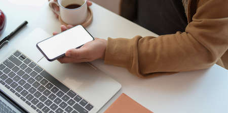 Cropped shot of young man holding blank screen smartphone while working on laptop and drinking a cup coffeeの写真素材