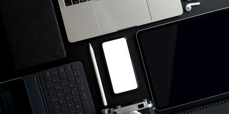 Top view of dark trendy workspace with blank screen smartphone and other electronics with office supplies on dark table backgroundの写真素材