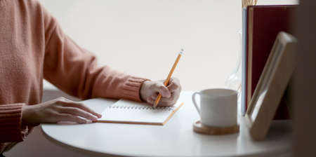 Close-up view of young female writing her idea on notebook while working on her project in comfortable roomの写真素材
