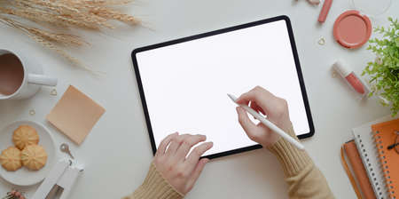 Top view of young female writing on blank screen tablet in warm beige feminine workspace concept with make up and office suppliesの写真素材