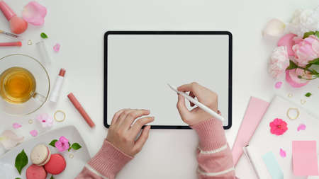 Top view of young girl writing on blank screen tablet in sweet pink feminine workspace with make up and office supplies の写真素材
