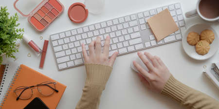 Top view of young female typing on keyboard in warm beige feminine workspace concept with make up...