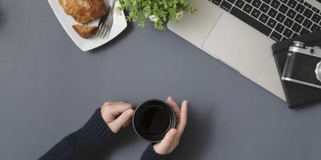 Top view of young female holding coffee cup in winter workspace with office supplies on grey desk backgroundの写真素材