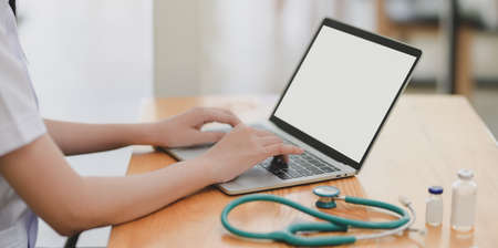 Close-up view of young female doctor examining the patient chart while using mock up laptop computer in her office room の写真素材