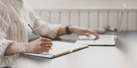 Close-up view of young passionate businesswoman typing on laptop computer and writing on notebook in office room の写真素材