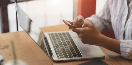 Cropped shot of young businesswoman holding smartphone while working on laptop computer in comfortable workplace の写真素材
