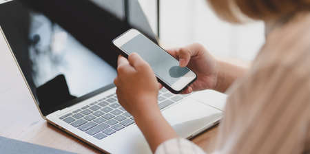 Cropped shot of young businesswoman holding smartphone while working on laptop computer in comfortable workplace の写真素材