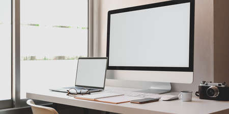 Blank screen desktop computer and laptop in modern workspace with office suppliesの写真素材