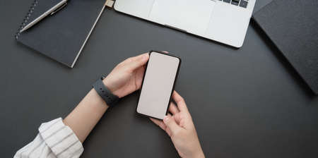 Overhead shot of young businesswoman holding blank screen smartphone in dark luxury workspace with office suppliesの写真素材