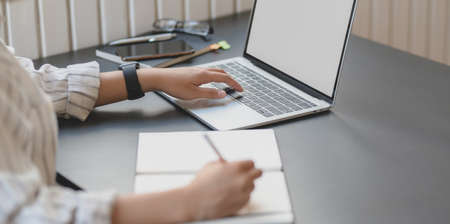Close-up view of young passionate businesswoman typing on laptop computer and writing on notebook in office roomの写真素材