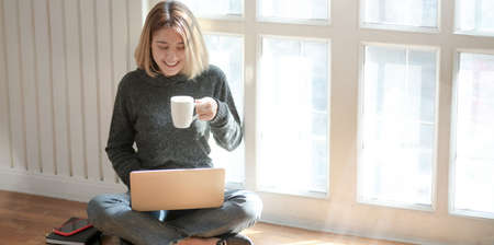 Portrait of young professional freelancer working on her project and drinking hot cocoa while sitting near the windows at her workplaceの写真素材