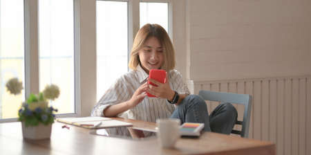 Young beautiful female designer looking at smartphone and sitting on the chair in her comfortable workspaceの写真素材