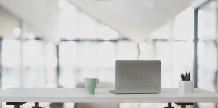 Laptop computer on white table and coffee cup in modern office room with blurred background