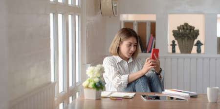 Portrait of young asian female designer looking at her smartphone while working in comfortable workspaceの写真素材