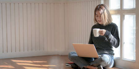 Portrait of young professional freelancer working on her project and drinking hot cocoa while sitting near the windows at her workplaceの写真素材