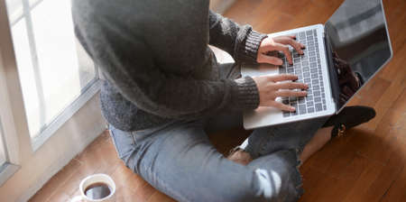 Close-up view of young female freelancer typing on laptop computer while sitting next to the windows with a cup of hot cocoaの写真素材