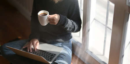 Close-up view of young freelancer working on her project and drinking hot cocoa while sitting near the windows at her workplaceの写真素材