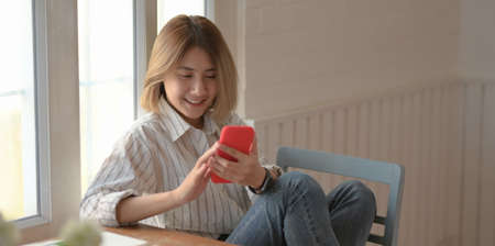 Young beautiful female designer looking at smartphone and sitting on the chair in her comfortable workspaceの写真素材