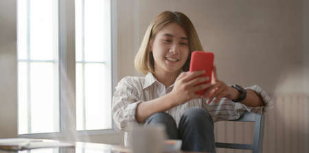 Young beautiful female designer looking at smartphone and sitting on the chair in her comfortable workspaceの写真素材