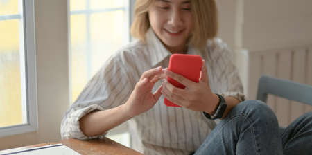 Young beautiful female designer looking at smartphone and sitting on the chair in her comfortable workspaceの写真素材
