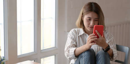 Portrait of young asian female designer looking at her smartphone while working in comfortable workspaceの写真素材