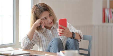 Young beautiful female designer looking at smartphone and sitting on the chair in her comfortable workspaceの写真素材