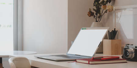 Minimal workspace with laptop computer, camera, office supplies and dry roses vase on white table