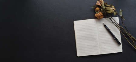 Top view of dark empty workspace with open notebook, pen and dry roses on black leather desk の写真素材