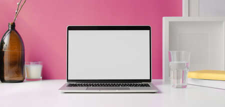 Modern workspace with open blank screen laptop computer with mock up frame and office supplies on white table and pink wall background の写真素材
