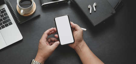 Top view of man holding blank screen smartphone in dark trendy workspace with office supplies 