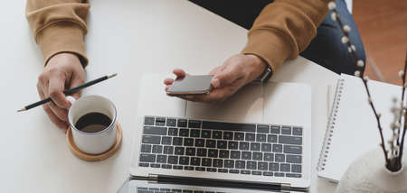Cropped shot of young man working while looking at smartphone and drinking a cup of coffee in comfortable workplaceの写真素材