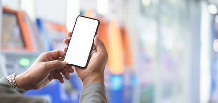 Cropped shot of young professional businessman holding blank screen smartphone with blurred ATM...