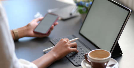 Close-up view of young businesswoman typing on digital tablet while using blank screen smartphone in...