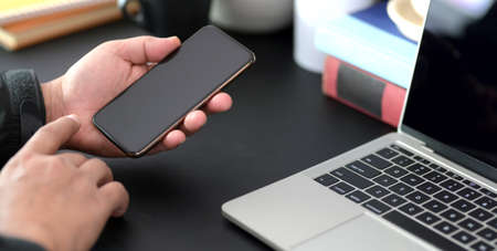 Cropped shot of young man holding smartphone in dark modern office room while working on his project...