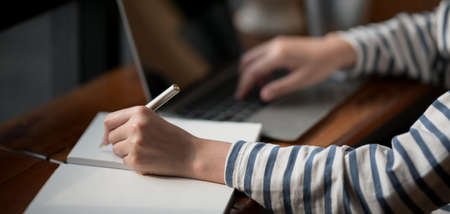 Close-up view of young woman working on her project while typing on laptop computer and writing the...