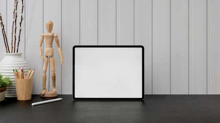 Close up view of artist workspace with blank screen tablet, wooden figure, coloured pencils and vase on black table with plank wall backgroundの写真素材