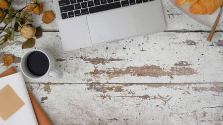 Top view of white vintage workspace with laptop, coffee cup, notebook, dry rose and   croissant on wooden table backgroundの写真素材