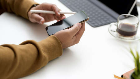 Cropped shot of professional artist holding on smartphone while working in simple workspace with tablet, stylus and coffee cup on white wooden tableの写真素材