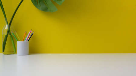 Cropped shot of colorful workspace with copy space, pencils and vase on white table with yellow wall background の写真素材