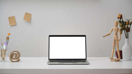Close up view of designer workspace with blank screen laptop, painting tools, wooden figure and decorations on white desk with white wall backgroundの写真素材