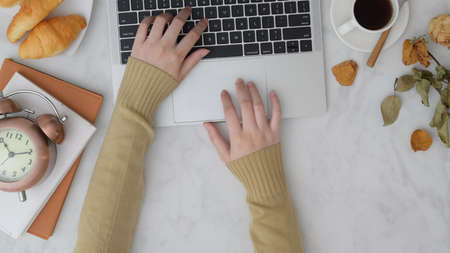Top view of female freelancer typing on laptop on vintage autumn workspace with breakfast, notebook and decorations on marble desk backgroundの写真素材