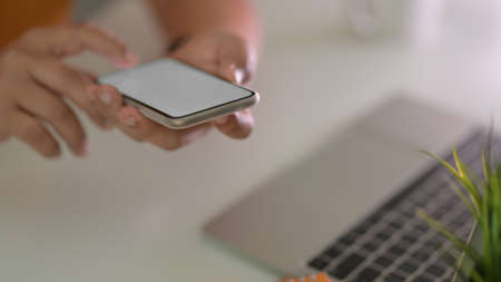 Cropped shot of male freelancer touching on blank screen smartphone in blurred workspace background