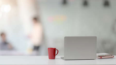 Close up view of simple workspace with laptop, notebooks, red coffee cup and copy space on white desk with blurred office room background の写真素材
