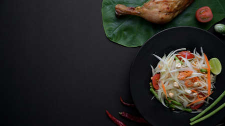 Top view of papaya salad on black plate, chicken grill on green taro and copy space with dark table backgroundの写真素材